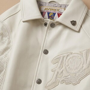 May include: Cream-colored leather jacket with a classic collar and silver-tone buttons. Features a perforated design on the left shoulder and an embroidered patch with the text "AVIREX" and "GOLD CLUB". A small metal emblem is on the collar.
