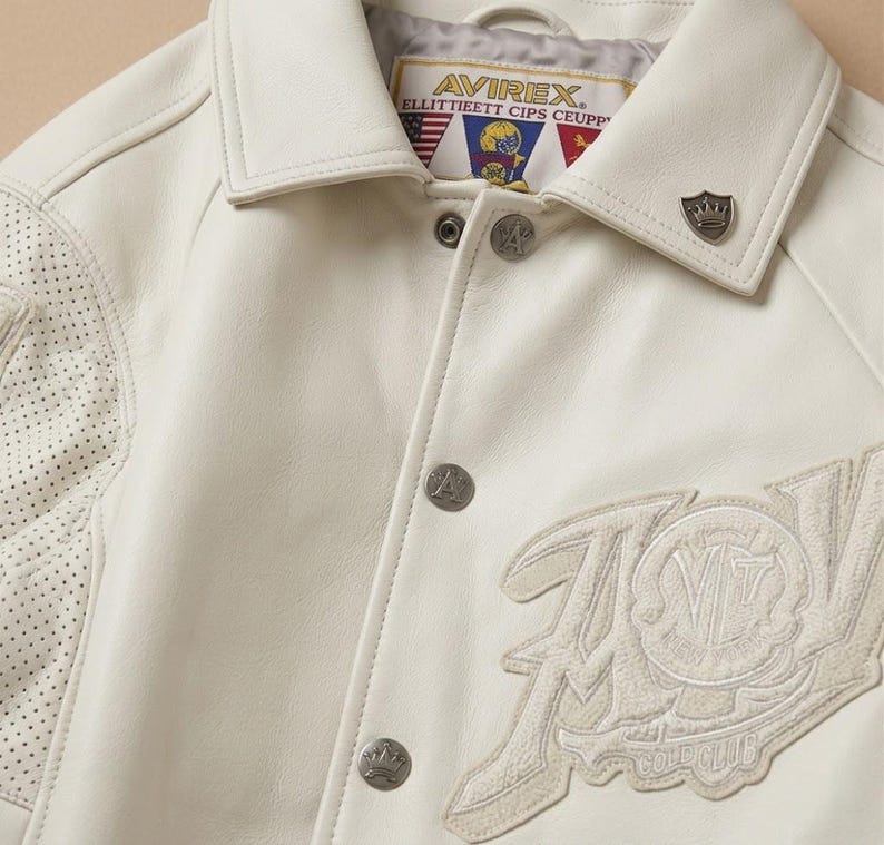 May include: Cream-colored leather jacket with a classic collar and silver-tone buttons. Features a perforated design on the left shoulder and an embroidered patch with the text "AVIREX" and "GOLD CLUB". A small metal emblem is on the collar.