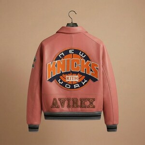 K&ouml;nnte beinhalten: Eine rosa Lederjacke mit dem Logo der New York Knicks auf dem R&uuml;cken. Die Jacke zeigt das Wort &bdquo;KNICKS&ldquo; in Orange und Blau, mit &bdquo;KITH&ldquo; und &bdquo;AVIREX&ldquo; ebenfalls sichtbar. Die Jacke hat einen grau-schwarzen gestreiften Saum und B&uuml;ndchen.