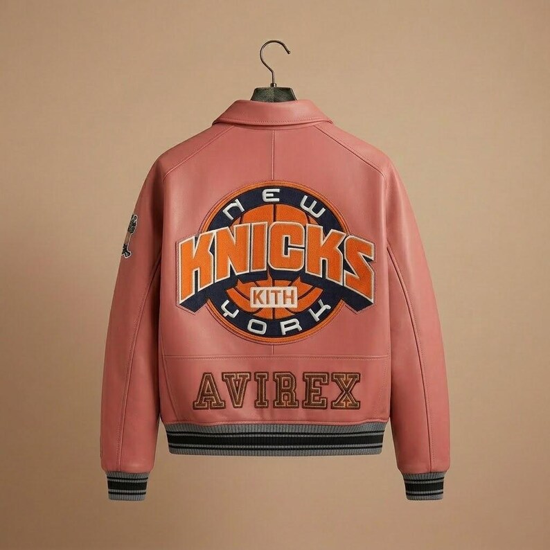 K&ouml;nnte beinhalten: Eine rosa Lederjacke mit dem Logo der New York Knicks auf dem R&uuml;cken. Die Jacke zeigt das Wort &bdquo;KNICKS&ldquo; in Orange und Blau, mit &bdquo;KITH&ldquo; und &bdquo;AVIREX&ldquo; ebenfalls sichtbar. Die Jacke hat einen grau-schwarzen gestreiften Saum und B&uuml;ndchen.