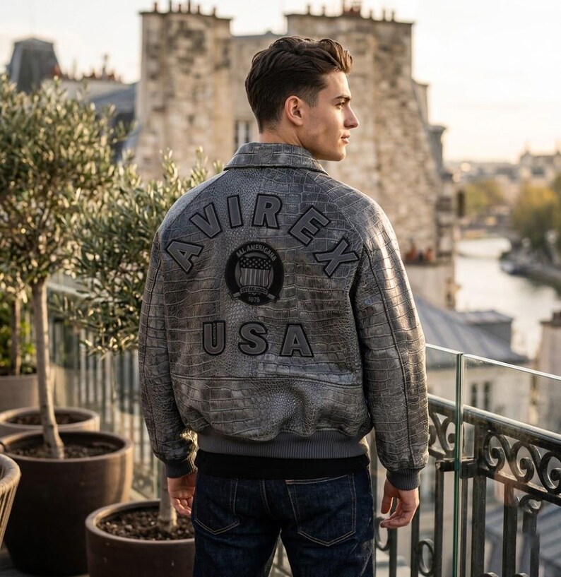 May include: A grey leather bomber jacket with a textured crocodile-skin pattern. The back features the black embroidered text AVIREX and USA, along with a circular patch displaying a flag graphic and the text ALL AMERICAN ESTABLISHED 1975.