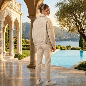 May include: A cream-colored jacket and matching pants set. The jacket features the word "Alma" and other emblems on the back. The person is wearing white sneakers. The background includes a pool, trees, and a scenic view.