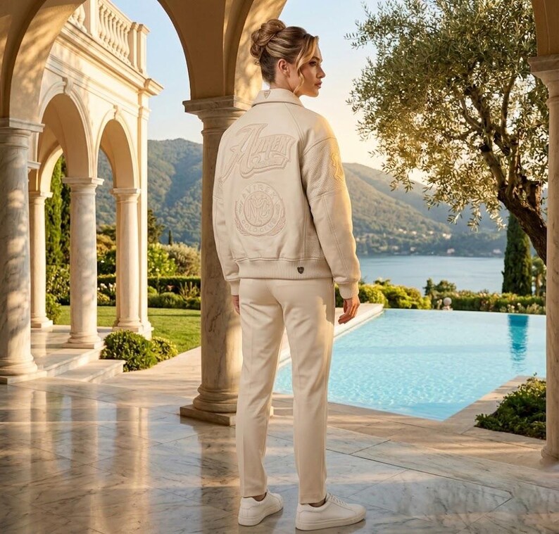 May include: A cream-colored jacket and matching pants set. The jacket features the word "Alma" and other emblems on the back. The person is wearing white sneakers. The background includes a pool, trees, and a scenic view.