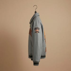 K&ouml;nnte beinhalten: Hellblaue Lederjacke mit schwarz-wei&szlig; gestreiften B&uuml;ndchen und Saum. Die Jacke hat Aufn&auml;her mit orangefarbenem und braunem Text und einer Basketballgrafik. Die Jacke h&auml;ngt an einem schwarzen Kleiderb&uuml;gel.