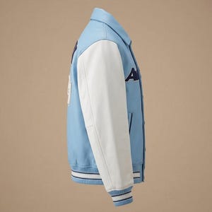 Puede incluir: Una chaqueta universitaria de color azul claro con mangas blancas, cuello en punta y cierre de botones. La chaqueta presenta letras azul oscuro en el pecho y un parche blanco en la espalda, con pu&ntilde;os y dobladillo de canal&eacute; a rayas.
