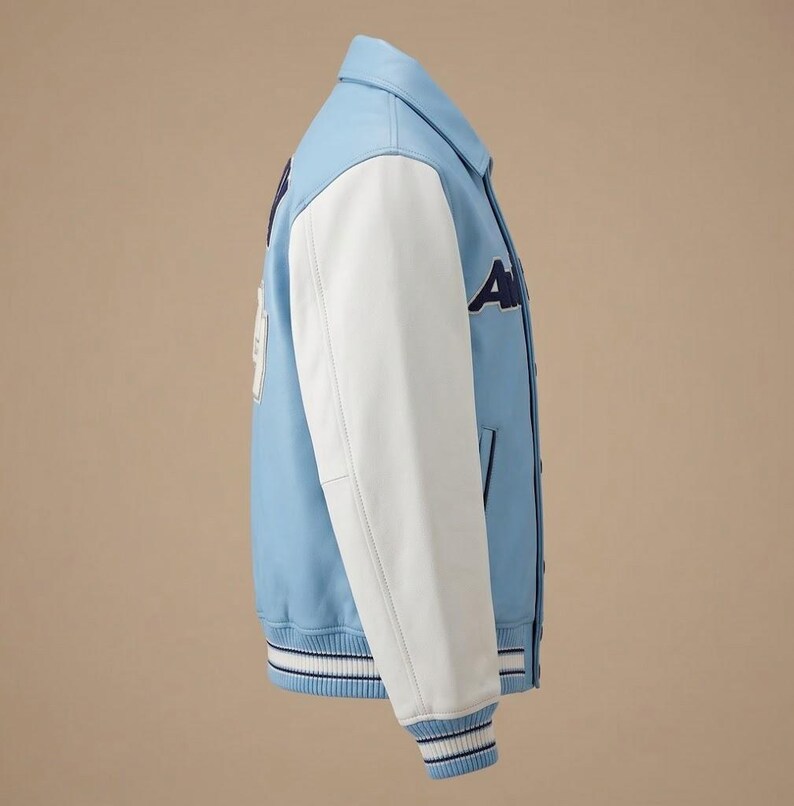 Puede incluir: Una chaqueta universitaria de color azul claro con mangas blancas, cuello en punta y cierre de botones. La chaqueta presenta letras azul oscuro en el pecho y un parche blanco en la espalda, con pu&ntilde;os y dobladillo de canal&eacute; a rayas.