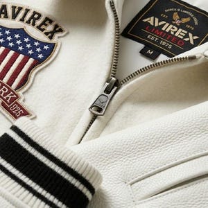 May include: An off-white varsity jacket featuring a textured leather body and a wool-blend chest panel. It displays an embroidered shield patch with the text AVIREX THE NEW YORK 1975 and a black interior label reading AVIREX LIMITED EST. 1975 M.