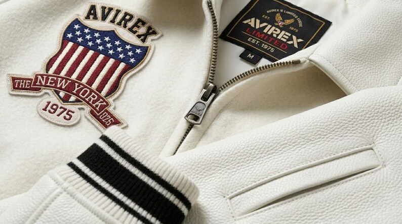 May include: An off-white varsity jacket featuring a textured leather body and a wool-blend chest panel. It displays an embroidered shield patch with the text AVIREX THE NEW YORK 1975 and a black interior label reading AVIREX LIMITED EST. 1975 M.