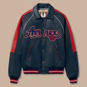 May include: Navy blue leather jacket with red accents on the shoulders and sleeves. The jacket features the word "Avirex" in a stylized font across the chest, with a red and blue emblem. White piping outlines the collar and shoulders.