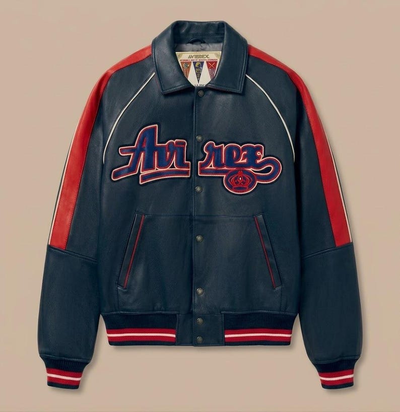 May include: Navy blue leather jacket with red accents on the shoulders and sleeves. The jacket features the word "Avirex" in a stylized font across the chest, with a red and blue emblem. White piping outlines the collar and shoulders.