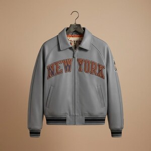K&ouml;nnte beinhalten: Graue Lederjacke mit dem Text "NEW YORK" in braunen und blauen Buchstaben auf der Brust. Die Jacke hat einen Kragen, einen Rei&szlig;verschluss und zwei Vordertaschen. Die Manschetten und der Bund haben schwarz-graue Streifen.