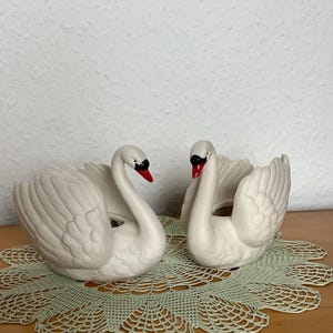 Può includere: Due statuine di cigno in ceramica bianca con occhi neri e becchi rossi. I cigni sono esposti su un centrino di pizzo verde chiaro. Le statuine hanno dettagliate texture di piume e sono su una superficie di legno.