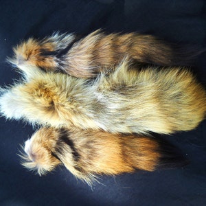 May include: Three fox tails in various shades of brown, white, and black. The tails are arranged in a pile on a black background.