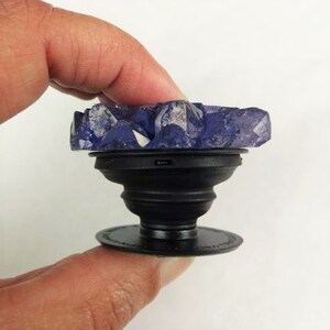 May include: A purple crystal phone grip with a black base. The crystal is shaped like a cluster of crystals.