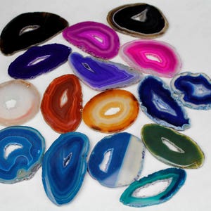 Puede incluir: Surtido de rodajas de ágata en varios colores, incluyendo azul, verde, naranja, rosa y blanco. Cada rodaja tiene un agujero natural en el centro, mostrando los intrincados patrones del ágata.