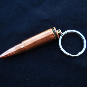 May include: A copper-colored bullet casing attached to a silver key ring. The bullet casing is about 2 inches long and has a pointed tip. The key ring is about 1 inch in diameter.
