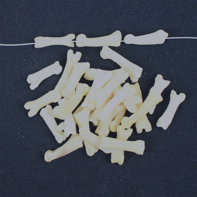 May include: Off-white bone-shaped beads, some strung on a thin white cord, others scattered on a dark blue surface. The beads vary in size and shape, resembling small bones. Suitable for jewellery making or crafting.