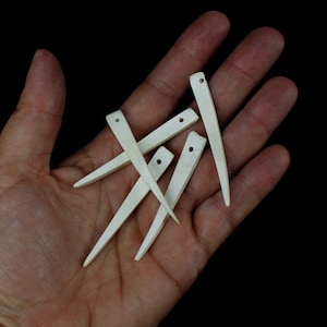 May include: Four white bone awls with holes at the top, arranged in a criss-cross pattern on a hand.