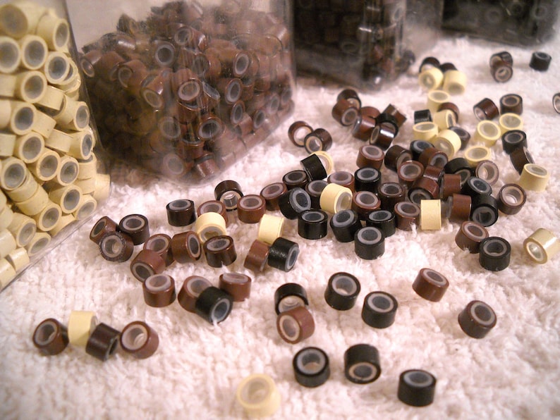 Puede incluir: Primer plano de una variedad de microanillos para extensiones de cabello en colores marr&oacute;n, negro y beige. Los anillos est&aacute;n esparcidos sobre una superficie blanca.