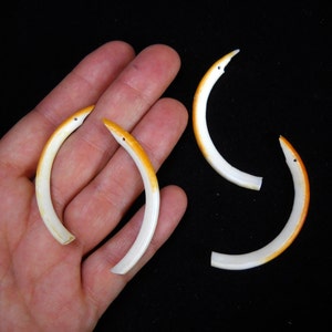 May include: Four curved, white and orange decorative pieces, resembling stylized birds or snakes, are displayed against a black background. The pieces have a curved shape with orange accents and small, dark eye details. The objects are arranged on a hand.