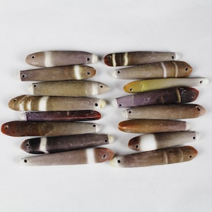 May include: A collection of 15 smooth, polished, and colorful sea shells with a hole drilled through each one. The shells are arranged in two rows, with the top row slightly offset from the bottom row. The shells are a variety of colors, including brown, white, and black.