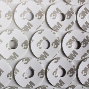 以下が含まれることがあります： 中央に穴が開いた白い丸い粘着ステッカーのシートのクローズアップ。各ステッカーには3Mのロゴが印刷されています。