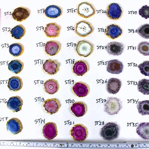 May include: Thirty-five agate slices with gold trim, arranged in rows on a white background. Each slice has a unique pattern of colors, including blue, purple, pink, and white.