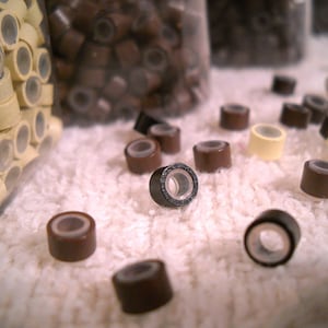 May include: A close-up of a variety of hair extension rings in brown and cream colors. The rings are scattered on a white fluffy surface.