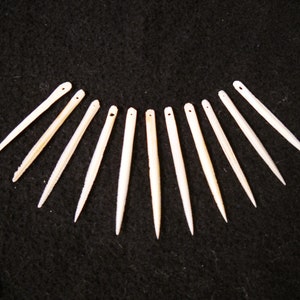 May include: A set of 11 white bone needles, arranged in a semi-circle on a black background. The needles are sharp and pointed, with a small hole at the top of each needle.