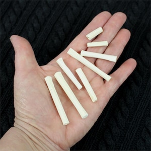 May include: A collection of eight white bone-like sticks of varying lengths, held in a person's hand. The sticks are smooth and have a natural, organic appearance.