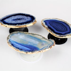 May include: Three blue and white agate phone grips with gold trim. The grips are shaped like a flat oval and have a raised, round base.