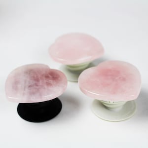 May include: Three pink heart-shaped phone grips made of rose quartz. Each grip has a different base, one black, one white, and one clear.