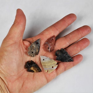 May include: Five arrowhead-shaped pendants made of agate. The pendants are in various colors, including brown, black, red, and white. Each pendant has two holes drilled through it.