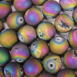 Puede incluir: Primer plano de una colección de cuentas redondas e iridiscentes. Las cuentas muestran un espectro de colores, incluyendo púrpura, dorado y azul. Algunas cuentas tienen una cavidad con formaciones de cristal brillantes. Cada cuenta tiene un agujero.