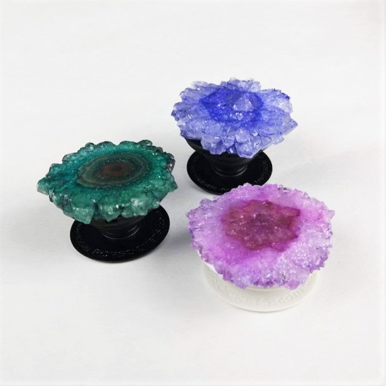 May include: Three phone grips with a crystal-like design. The grips are in shades of purple, green, and pink. Each grip has a round base and a raised crystal design.
