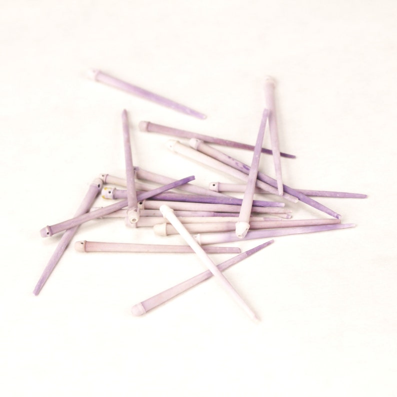 May include: A pile of approximately 20 purple and white slender sticks, possibly made of wood or bone, on a white background.