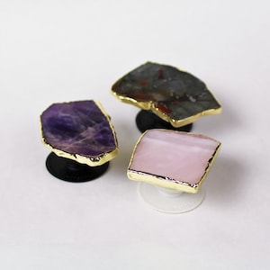 May include: Three phone grips with different coloured stone tops. The first grip has a purple amethyst stone, the second has a pink rose quartz stone, and the third has a grey and red agate stone. All three grips have gold trim around the stone and a black base.