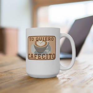 May include: White coffee mug with a handle. The mug features a rectangular design with the words "YO QUIERO CAFECITO" and an illustration of a coffee cup with latte art. The mug is on a wooden surface.