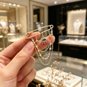 May include: Gold-toned safety pin brooch with dangling charms. The brooch features an eye pendant with black and white details, star-shaped charms, and delicate chain accents. Small pearl-like beads add to the design. The brooch is held up against a blurred background.
