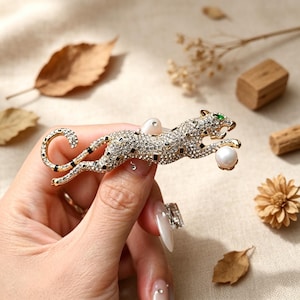 May include: A gold-toned brooch in the shape of a leaping leopard, encrusted with clear crystals and black accents. The leopard holds a faux pearl in its paw and has green eyes. The brooch is approximately 3 inches long.