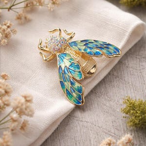 May include: A gold-toned bee brooch with iridescent rhinestones and enamel wings in shades of blue and green. The brooch is set on a white cloth, with dried flowers in the background.