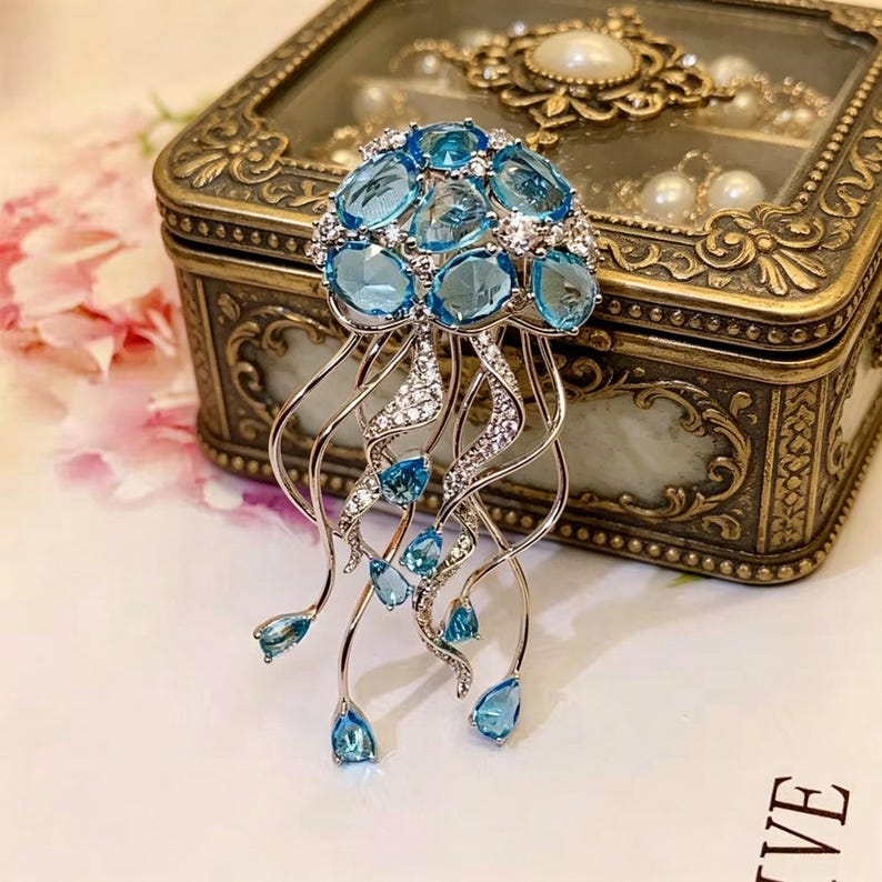 Puede incluir: Broche plateado con forma de medusa, con un grupo de piedras preciosas azules claras de forma ovalada en la parte superior. El broche tiene zarcillos curvos y fluidos adornados con cristales transparentes y gemas azules en forma de l&aacute;grima. En el fondo, una caja de joyer&iacute;a decorativa.