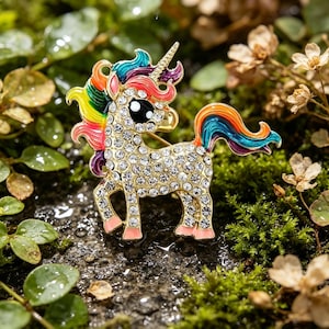 May include: A gold-toned unicorn brooch, encrusted with clear crystals, rests on a mossy surface. The unicorn features a rainbow mane and tail, black eyes, and pink hooves. The brooch is a whimsical accessory.