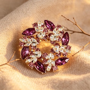 May include: A gold-toned brooch with a circular design. The brooch features large, deep purple marquise-cut crystals and smaller pink and clear crystals, arranged in a floral pattern. The brooch is set against a neutral fabric background.