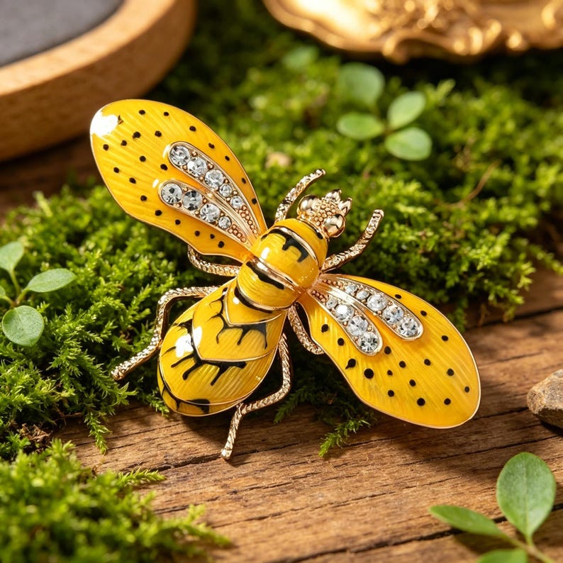 May include: A yellow and black bee-shaped brooch with rhinestone accents. The brooch has a gold-tone metal frame and is set against a backdrop of green moss and wood. The bee's wings are adorned with small black dots.