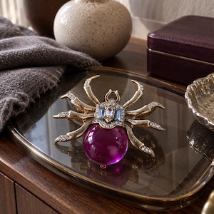 May include: A spider-shaped brooch featuring a large, round, translucent purple stone as the abdomen. The body and eight legs are crafted from gold-toned metal, accented with small clear rhinestones and rectangular light blue crystals.