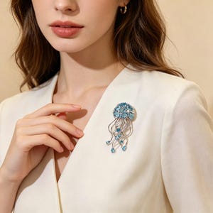 Puede incluir: Broche plateado con forma de medusa. La cabeza de la medusa est&aacute; adornada con piedras preciosas azul claro, con hebras plateadas y piedras en forma de l&aacute;grima. El broche est&aacute; prendido a una blazer blanca.