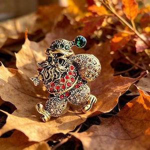 Pode incluir: Broche dourado do Pai Natal com chapéu verde e detalhes vermelhos. O broche apresenta um design detalhado com um saco de presentes sobre o ombro, sobre um fundo de folhas de outono. O broche tem cerca de 5 cm de altura.