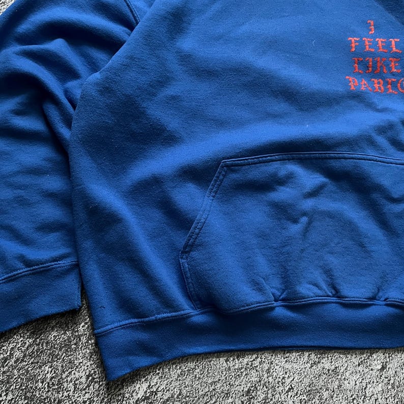 May include: A blue hoodie with a front pocket and the text "I FEEL LIKE PABLO" in orange. The hoodie is made of a soft material and has a drawstring hood.