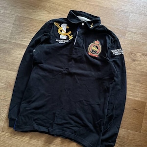May include: A black long-sleeve polo shirt featuring a white number one and gold embroidered horseshoe logo with the text MERCER CLUB MMVIII. The chest displays a circular crest with a horse and rider, and the sleeve has a patch reading MERCER COMPETITION WINTER M.
